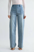A woman modeling a pair of high-waisted, wide-leg front seam medium blue jeans with size zipper and a light pink off-the-shoulder long-sleeve top, standing against a plain light gray studio background.