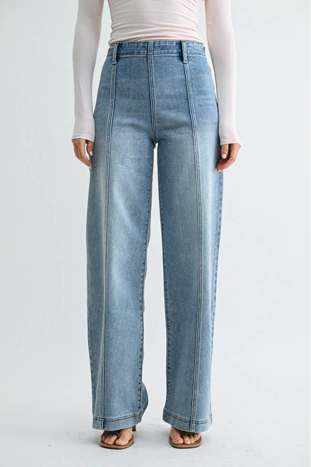 A woman modeling a pair of high-waisted, wide-leg front seam medium blue jeans with size zipper and a light pink off-the-shoulder long-sleeve top, standing against a plain light gray studio background.