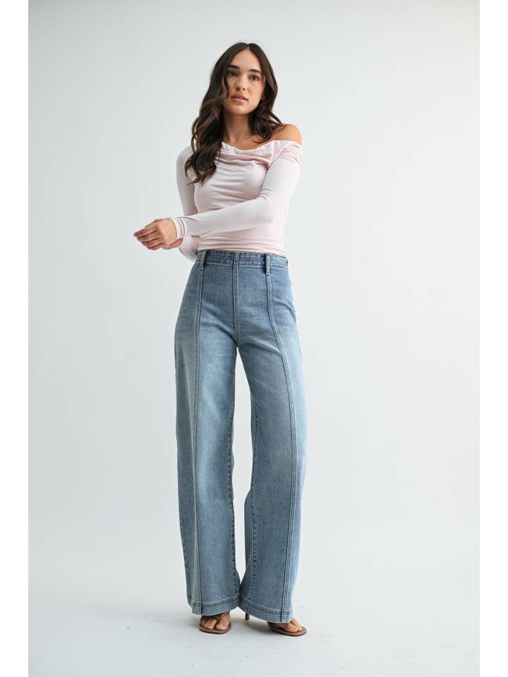 A woman modeling a pair of high-waisted, wide-leg front seam medium blue jeans with size zipper and a light pink off-the-shoulder long-sleeve top, standing against a plain light gray studio background.