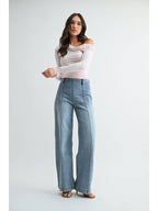 A woman modeling a pair of high-waisted, wide-leg front seam medium blue jeans with size zipper and a light pink off-the-shoulder long-sleeve top, standing against a plain light gray studio background.