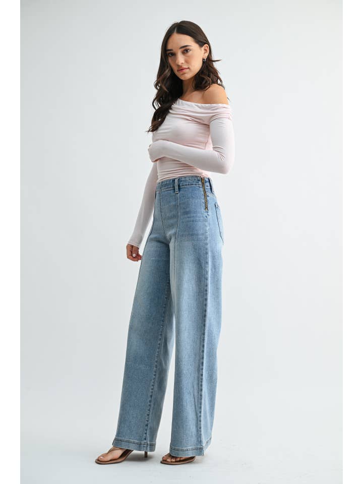 A woman modeling a pair of high-waisted, wide-leg front seam medium blue jeans with a unique side zipper detail, paired with a light pink off-the-shoulder long-sleeve top and brown heeled sandals, against a plain light gray studio background.