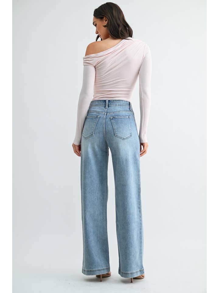 Back view of a woman wearing a light pink off-the-shoulder long-sleeve top and medium blue wide-leg jeans against a plain light gray studio background.