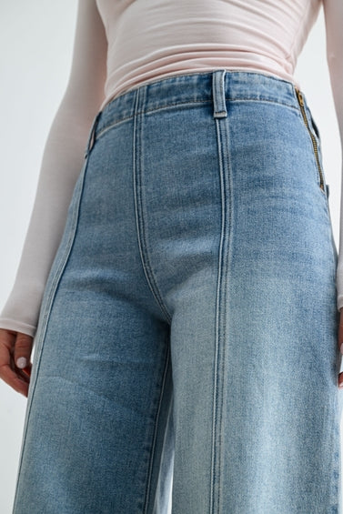 Close-up of a woman wearing medium blue wide-leg denim jeans with a front seam and pale pink long-sleeve top, shown from the waist to mid-thigh against a plain light background.