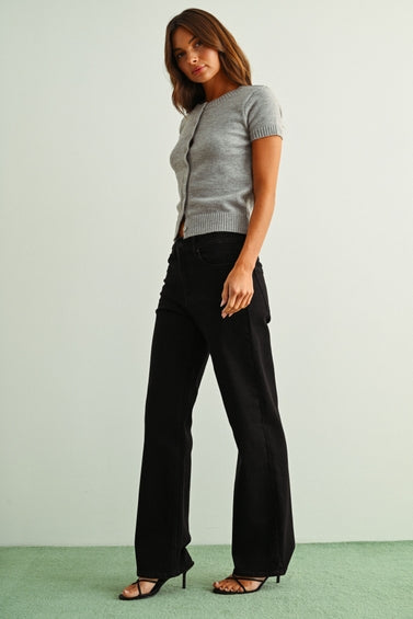 A woman wearing black straight jeans, a light gray sleeveless knit top, and brown sandals, standing against a light gray wall and green floor.