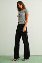 A woman wearing black straight jeans, a light gray sleeveless knit top, and brown sandals, standing against a light gray wall and green floor.
