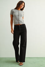 A woman wearing black straight jeans, a light gray sleeveless knit top, and brown sandals, standing against a light gray wall and green floor.