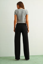 A woman shown from the back wearing black straight jeans, a light gray sleeveless knit top, and brown sandals, standing against a light gray wall and green floor.