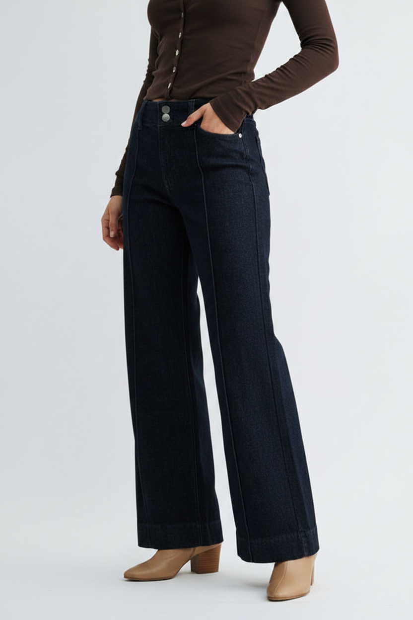 A model wearing dark wash wide-leg jeans, a brown long-sleeve top, and tan heeled shoes, standing against a light gray studio background.