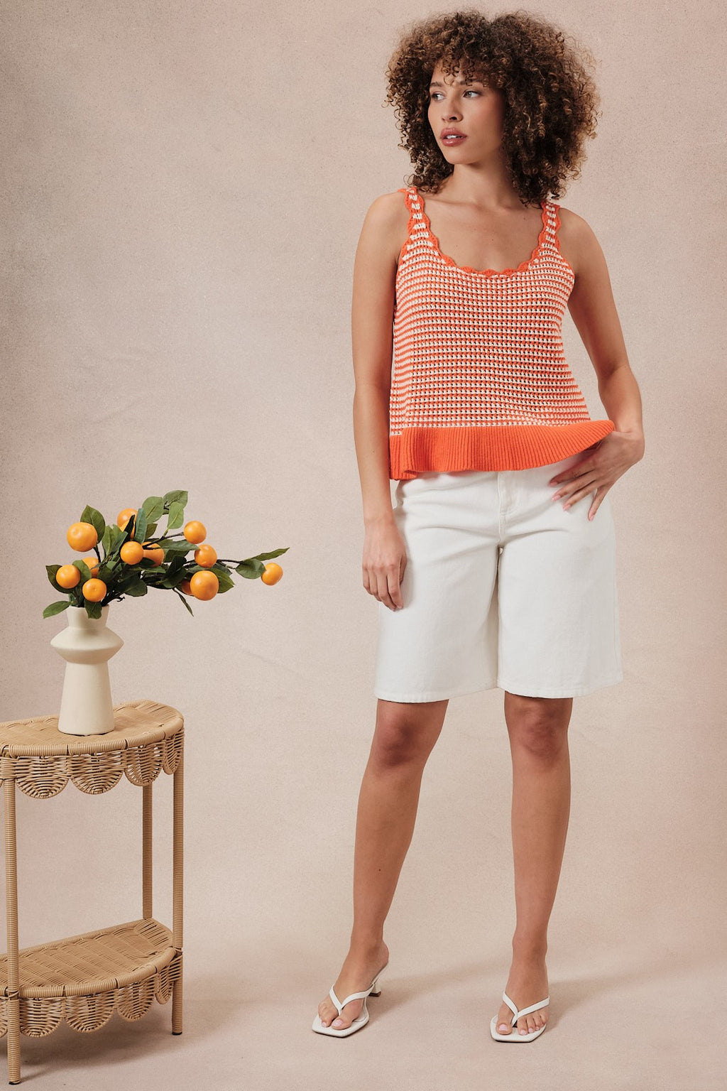 Woman wearing an orange and white  striped crochet knit tank top and white shorts against a beige background