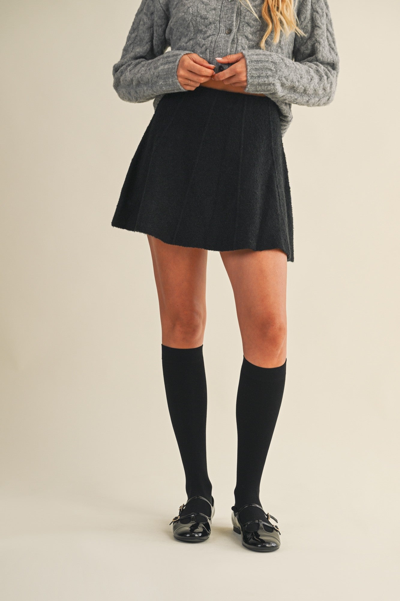 Woman wearing a black knit mini skirt, gray sweater, and black knee-high socks on a beige background