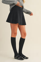 Woman wearing a black knit mini skirt, gray sweater, and black knee-high socks on a beige background