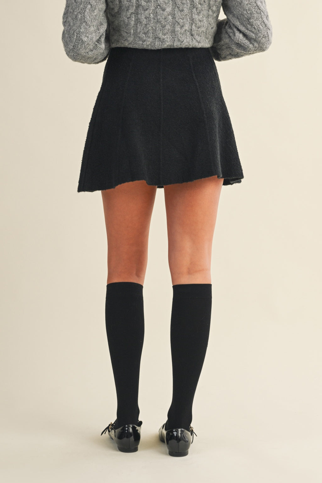 Back view of a woman wearing a black knit mini skirt, gray sweater, and black knee-high socks on a beige background