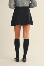 Back view of a woman wearing a black knit mini skirt, gray sweater, and black knee-high socks on a beige background