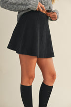 Woman wearing a black knit mini skirt, gray sweater, and black knee-high socks on a beige background
