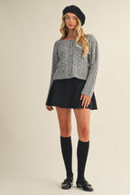 Woman wearing a black knit mini skirt, gray sweater, and black knee-high socks on a beige background