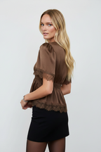 A blonde model wearing a brown short-sleeve blouse with lace trim and a black mini skirt, photographed against a plain light gray studio background.