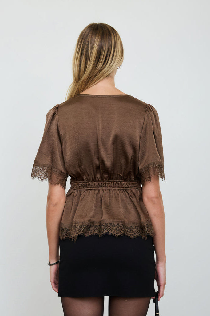 A woman shown from the back wearing a brown short-sleeve top with lace trim and a black skirt, photographed against a plain light gray studio background.