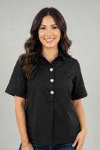 A woman modeling a black short sleeve blouse with gold tone buttons and front pleat against a white background.
