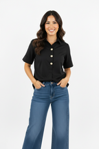 A woman modeling a black short sleeve blouse with gold tone buttons and front pleat tucked in jeans and against a white background.