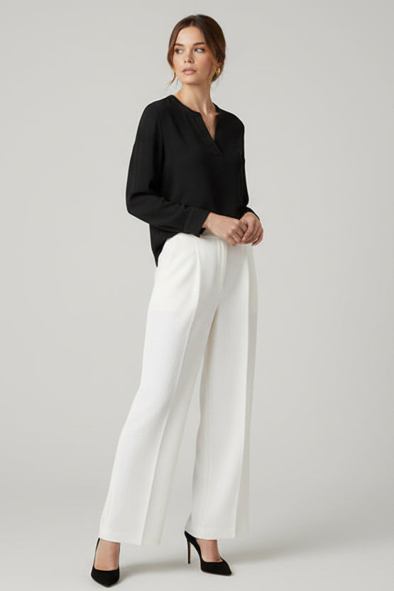 Woman wearing a black long sleeve textured top with white pants against a plain background