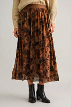 A woman shown wearing a mocha floral maxi skirt, a beige sweater, and brown ankle boots, standing against a solid dark green backdrop.