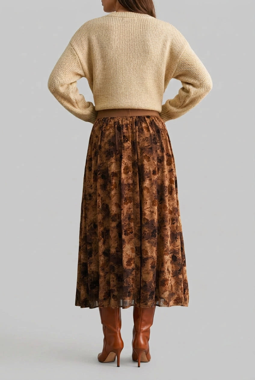 A woman shown from the back wearing a mocha floral maxi skirt, a beige sweater, and brown ankle boots, standing against a solid dark green backdrop.