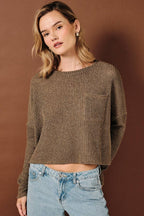 A woman wearing a brown ribbed knit long-sleeve crop top sweater with a chest pocket, paired with light blue jeans, standing against a solid brown studio background.