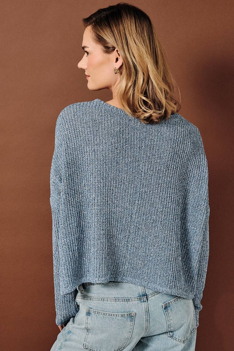 A woman shown from the back wearing a textured denim blue long-sleeve knit sweater with a chest pocket, paired with light-wash blue jeans, standing against a solid brown studio backdrop.