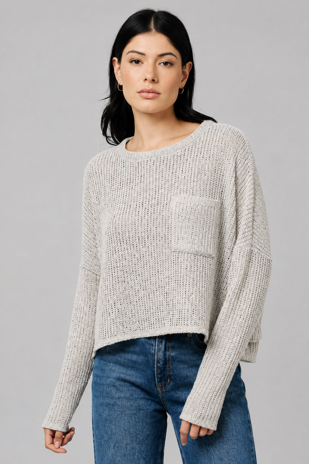 A woman wearing a natural color textured knit long-sleeve top with a chest pocket, paired with blue jeans, standing against a neutral beige wall.