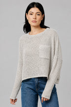A woman wearing a natural color textured knit long-sleeve top with a chest pocket, paired with blue jeans, standing against a neutral beige wall.