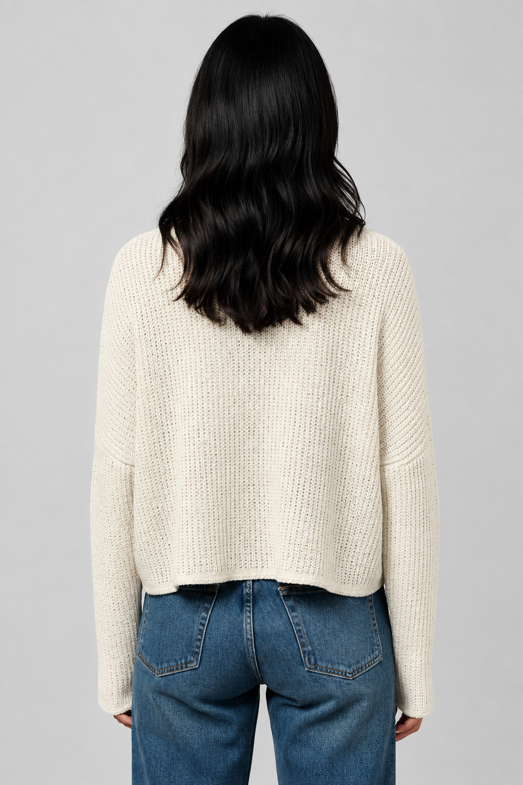 Back view of a woman wearing a natural color textured knit long-sleeve top, paired with blue jeans, standing against a neutral beige wall.