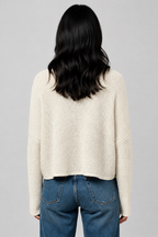 Back view of a woman wearing a natural color textured knit long-sleeve top, paired with blue jeans, standing against a neutral beige wall.