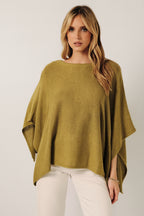 A woman wearing a loose, green knit layering sweater cape with white pants, standing against a soft white curtain backdrop.