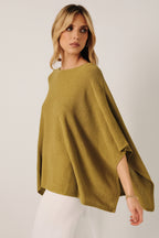 A woman wearing a loose, green knit layering sweater cape with white pants, standing against a soft white curtain backdrop.