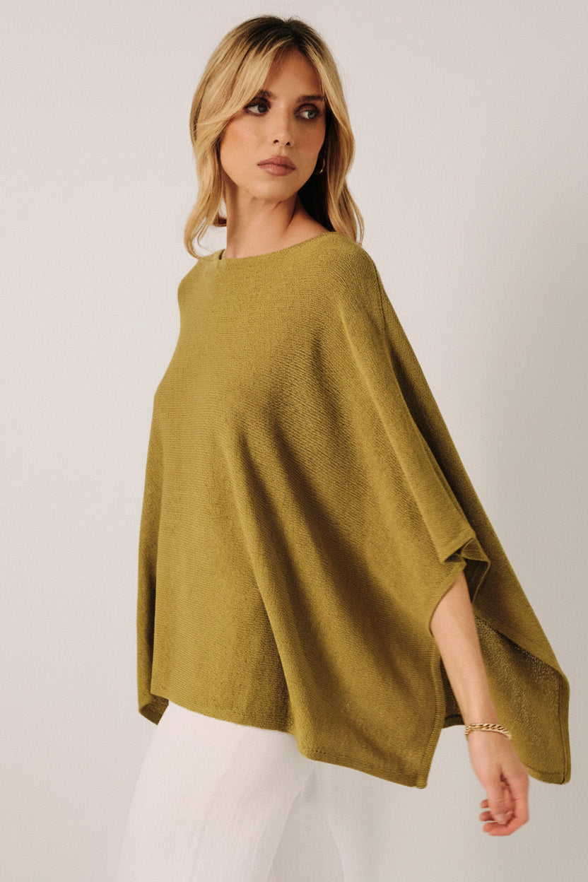 A woman wearing a loose, green knit layering sweater cape with white pants, standing against a soft white curtain backdrop.