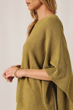 A woman wearing a loose, green knit layering sweater cape against a soft white curtain backdrop.