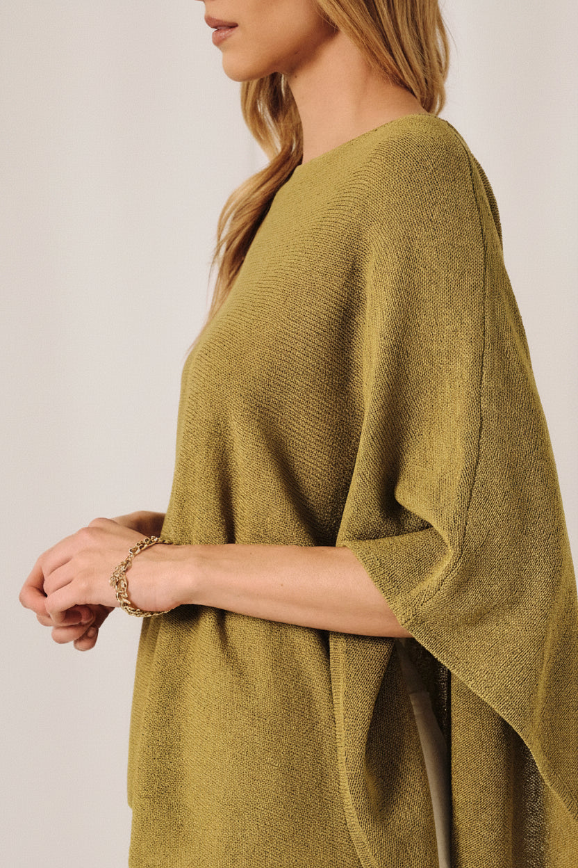 A woman wearing a loose, green knit layering sweater cape against a soft white curtain backdrop.
