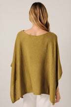 Back view of a woman wearing a loose, green knit layering sweater cape with white pants, standing against a soft white curtain backdrop.