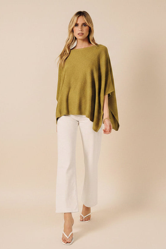 Woman wearing a green sweater cape and white pants against a white background