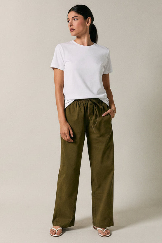 A woman modeling olive green straight-leg drawstring pants with a white t-shirt, standing against a solid beige studio background.