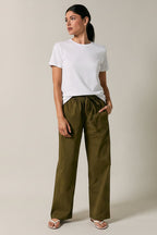 A woman modeling olive green straight-leg drawstring pants with a white t-shirt, standing against a solid beige studio background.