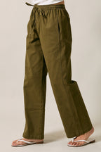 A woman modeling olive green straight-leg drawstring pants standing against a solid beige studio background.