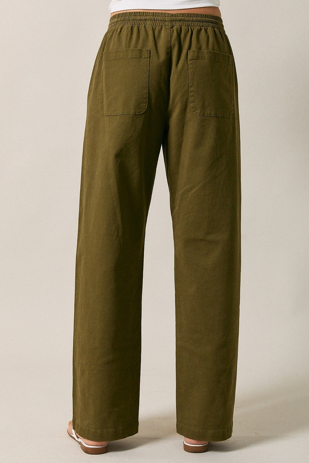 Back view of a woman modeling olive green straight-leg drawstring pants standing against a solid beige studio background.