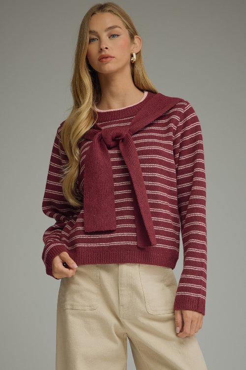 A woman modeling a berry and pink striped knit sweater with a draped shoulder accent at the neckline, paired with light beige trousers, against a solid gray studio background.