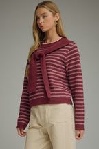 A woman modeling a berry and pink striped knit sweater with a draped shoulder accent at the neckline, paired with light beige trousers, against a solid gray studio background.