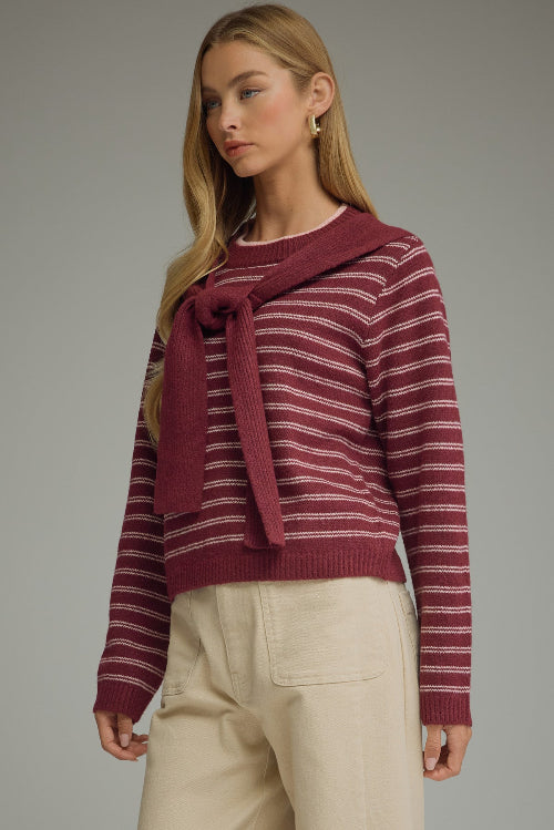 A woman modeling a berry and pink striped knit sweater with a draped shoulder accent at the neckline, paired with light beige trousers, against a solid gray studio background.