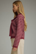 A woman modeling a berry and pink striped knit sweater with a draped shoulder accent at the neckline, paired with light beige trousers, against a solid gray studio background.