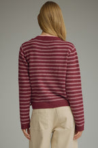 Back view of a woman modeling a berry and pink striped knit sweater with a draped shoulder accent at the neckline, paired with light beige trousers, against a solid gray studio background.