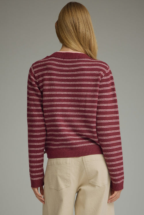 Back view of a woman modeling a berry and pink striped knit sweater with a draped shoulder accent at the neckline, paired with light beige trousers, against a solid gray studio background.