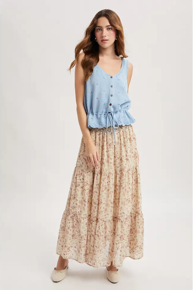 Woman wearing a sky blue sleeveless knitted tank top with buttons and print skirt on a plain background